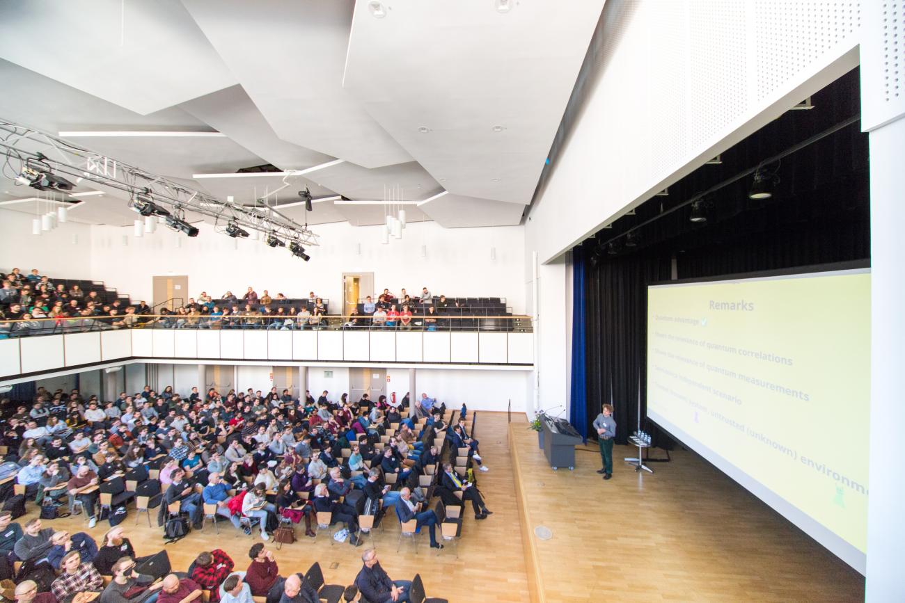 Links ein sitzendes Publikum, rechts eine Bühne mit Vortragendem