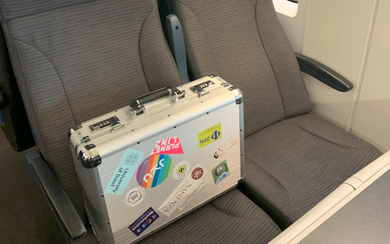A silver suitcase with many stickers sitting in a train seat